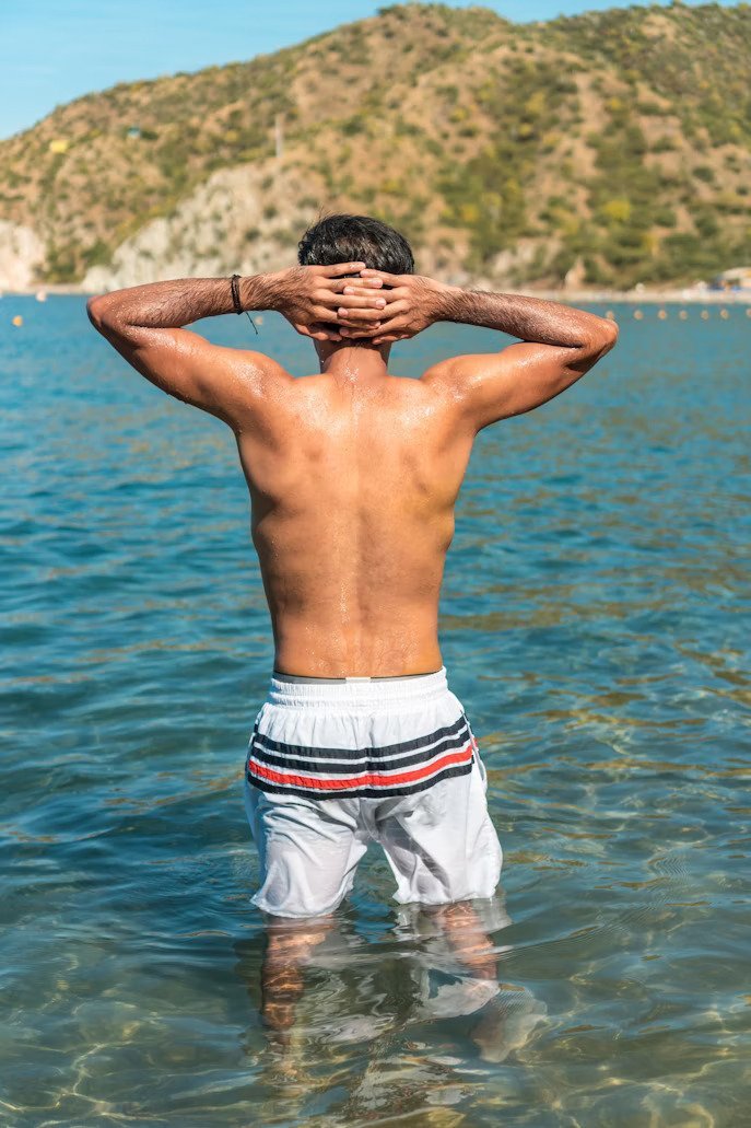 Before: Before and after AI preview of a wider back and lats on a man standing in the sea with hands on his head, made in Buldy