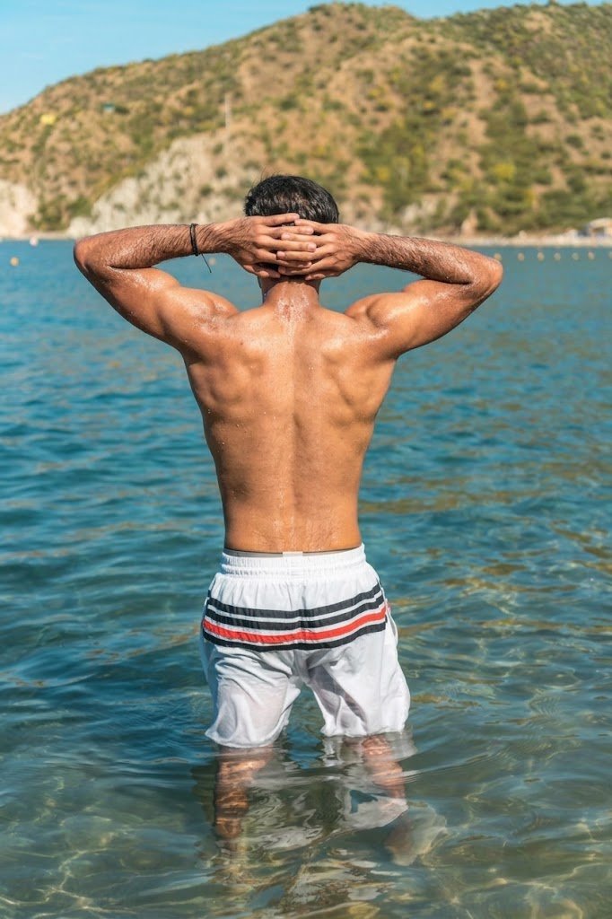 After: Before and after AI preview of rounder, fuller shoulders and rear delts on a man standing in the sea with hands on his head, made in Buldy